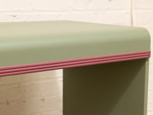 Load image into Gallery viewer, A close-up of a table corner showing a muted sage green top and base with a contrasting dusty rose pink beaded edge trim, set against a white brick wall background.
