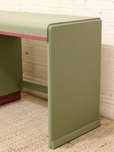 Load image into Gallery viewer, A close-up of a painted wooden desk or console table leg and side panel in a muted sage green, with a pink edge stripe and a dark red base visible against a white brick wall and beige rug.
