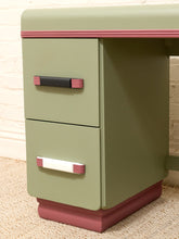 Load image into Gallery viewer, A close-up of a vintage-style two-drawer desk or cabinet in muted sage green with dusty rose trim and matching drawer pulls, shown against a light brick wall and carpet.
