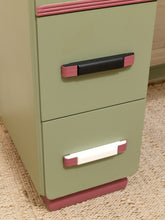 Load image into Gallery viewer, A close-up of a vintage-style two-drawer filing cabinet in sage green with pink trim and modern black and white handles, resting on a beige woven rug.

