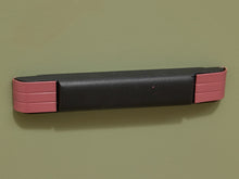 Load image into Gallery viewer, A black rectangular handlebar grip cover with pink end caps, shown mounted on a plain olive-green wall.
