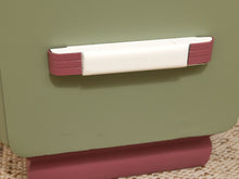 Load image into Gallery viewer, A close-up of a vintage-style drawer or cabinet handle with a white center and dusty rose pink end caps, mounted on a muted sage green wooden surface.
