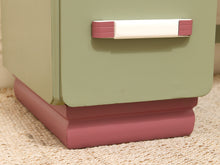 Load image into Gallery viewer, A close-up of a vintage-style nightstand or cabinet corner, featuring a sage green upper body, a dusty rose base, and a matching dusty rose and white drawer handle.
