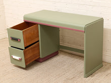 Load image into Gallery viewer, A mid-century modern writing desk in sage green with pink trim and two open drawers showing a wood interior, set against a white brick wall and beige carpet.
