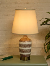 Load image into Gallery viewer, A table lamp with a warm white drum shade and a speckled ceramic base in brown, white, and gold tones, sitting on a green side table against a white brick wall with a plant on the right.
