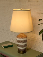 Load image into Gallery viewer, A table lamp with a warm white fabric drum shade and a textured ceramic base featuring horizontal bands of white, brown, and gold glitter, sitting on a green side table against a white brick wall.
