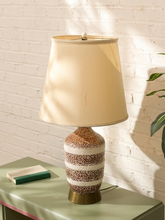 Load image into Gallery viewer, A table lamp with a beige fabric drum shade and a speckled ceramic base in brown, cream, and gold tones, sitting on a green side table against a white brick wall.
