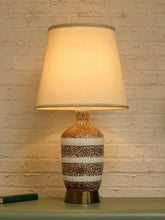 Load image into Gallery viewer, A table lamp with a warm white tapered fabric shade and a speckled ceramic base in brown, white, and gold tones, sitting on a tabletop against a white brick wall.
