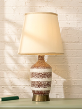 Load image into Gallery viewer, A table lamp with a warm cream fabric shade and a speckled ceramic base in brown, white, and gold tones, sitting on a light green surface against a white brick wall.

