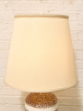 Load image into Gallery viewer, A cream-colored fabric lampshade with a flared silhouette, mounted on a speckled ceramic base against a white painted brick wall.
