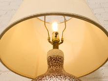 Load image into Gallery viewer, A close-up of a table lamp with a large warm cream fabric shade, a brass socket, and a speckled ceramic base against a white brick wall background.
