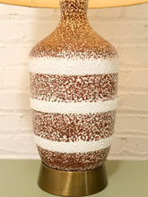 Load image into Gallery viewer, A vintage-style table lamp with a brown and white speckled ceramic vase body and a brass-colored metal base, set against a white brick wall.
