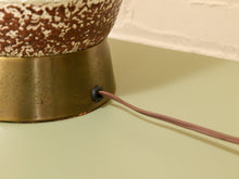 Load image into Gallery viewer, Close-up of a table lamp base with a speckled brown and cream ceramic upper section, a brushed brass-colored metal base, and a brown power cord exiting through a side socket.

