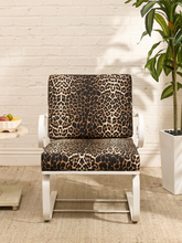 Load image into Gallery viewer, A white-framed accent chair with leopard-print cushions, set against a white brick wall with a potted plant and a small side table in the background.

