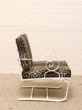 Load image into Gallery viewer, A white metal rocking chair with leopard-print cushions, set against a white brick wall and a beige woven rug.

