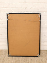 Load image into Gallery viewer, The back of a framed picture with a brown cardboard backing and a black metal frame, leaning against a white brick wall on a beige carpet.
