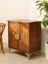 Load image into Gallery viewer, A mid-century modern wooden cabinet with sunburst carved doors and brass legs, set against a white brick wall with a potted plant nearby.
