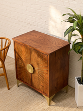 Load image into Gallery viewer, A mid-century modern wooden cabinet with a sunburst brass front detail and brass legs, shown in a styled room setting next to a plant and chair.
