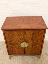 Load image into Gallery viewer, Mid-century modern wooden cabinet with a warm brown wood finish, brass sunburst door details, and brass legs, set against a white brick wall and woven rug.
