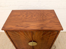 Load image into Gallery viewer, A mid-century modern wooden side table or cabinet with a warm brown wood grain finish, featuring a circular brass pull handle and radial slat detailing on the front doors, set against a white brick wall and beige carpet.
