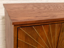 Load image into Gallery viewer, Close-up of a mid-century modern wooden credenza or sideboard cabinet, showing the warm brown wood grain top and two doors with a gold sunburst inlay pattern, set against a white brick wall background.
