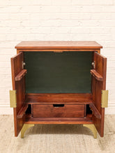 Load image into Gallery viewer, A mid-century modern wooden cabinet with open doors, showcasing a dark green interior lining, a lower drawer, and brass hardware, set against a white brick wall and beige rug.
