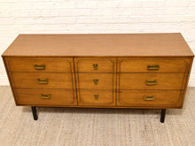Load image into Gallery viewer, A mid-century modern wooden dresser with six drawers, featuring a warm medium-brown wood finish, brass handles, and black legs, set against a white brick wall and beige carpet.
