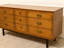 Load image into Gallery viewer, A mid-century modern wooden dresser with six drawers, featuring a warm honey-amber wood finish, brass handles, and black legs, set against a white brick wall and beige carpet.
