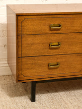 Load image into Gallery viewer, A close-up of a mid-century modern wooden three-drawer dresser with brass handles and black legs, set against a white brick wall and beige carpet.
