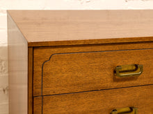 Load image into Gallery viewer, Close-up of a wooden chest of drawers with a warm medium-brown finish and brass handles, set against a white brick wall.
