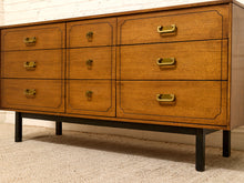 Load image into Gallery viewer, A mid-century modern wooden dresser with six drawers, featuring a warm medium-brown wood grain finish, brass handles, and black metal legs, set against a white brick wall and beige carpet.
