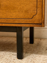 Load image into Gallery viewer, Close-up detail of a mid-century modern wooden cabinet or side table, showing a warm medium-brown wood grain top with a black painted lower frame and legs, standing on a woven beige rug.
