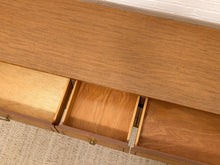 Load image into Gallery viewer, Close-up of a wooden furniture piece with multiple drawers, showing the warm medium-brown wood grain and brass drawer pulls, set against a light textured carpet and white brick wall.

