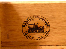Load image into Gallery viewer, Close-up of a wooden surface with a circular black laser-etched logo reading 'BASSETT FURNITURE INDUSTRIES, INC.' around a covered wagon illustration.
