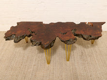 Load image into Gallery viewer, A rustic live-edge wood coffee table with a dark brown and black bark-veined top, supported by four slim gold metal hairpin legs, set on a beige woven rug against a white brick wall.
