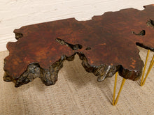 Load image into Gallery viewer, A rustic live-edge wooden coffee table with a rich dark brown and reddish-brown wood grain top, supported by three slim gold metal hairpin legs, set on a woven jute rug against a white brick wall.
