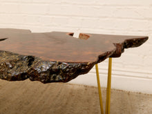 Load image into Gallery viewer, A rustic live-edge wooden table with a dark brown and black textured top and slim gold metal hairpin legs, shown against a white brick wall and beige rug.
