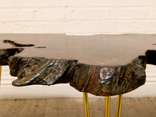 Load image into Gallery viewer, A rustic live-edge wooden table with a dark brown and black bark-look top, supported by slim gold metal legs, set against a white brick wall background.

