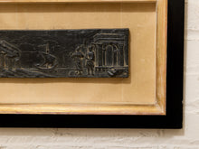Load image into Gallery viewer, Close-up of a framed art piece featuring a dark carved relief plaque with classical architectural and human figures, mounted on a beige mat within a black and natural wood frame, set against a white brick wall.
