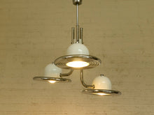 Load image into Gallery viewer, A three-light chandelier with a polished silver metal frame and white glass globes, shown hanging against a beige brick wall.
