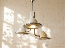Load image into Gallery viewer, A modern three-light pendant chandelier with cream-colored dome shades and polished silver/chrome metal accents, hanging against a white brick wall with soft window shadow patterns.
