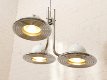 Load image into Gallery viewer, A three-light industrial ceiling fixture with polished chrome curved arms and white dome shades, shown illuminated against a white brick wall.

