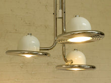 Load image into Gallery viewer, A mid-century modern three-light chandelier with glossy white dome-shaped glass shades and polished chrome metal arms, shown illuminated against a light brick wall.
