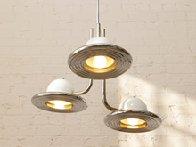 Load image into Gallery viewer, A modern three-light pendant chandelier with brushed silver metal shades and white dome-shaped diffusers, shown lit against a white brick wall.
