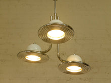 Load image into Gallery viewer, A three-light pendant chandelier with polished chrome metal arms and circular ribbed shades, each holding a white dome-shaped glass shade that glows with warm light, hanging against a beige brick wall.
