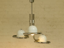 Load image into Gallery viewer, A modern three-light chandelier with a polished chrome metal frame and white glass globes, hanging against a textured beige brick wall.
