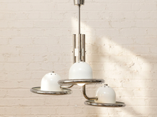 Load image into Gallery viewer, A modern three-light pendant chandelier with white dome-shaped glass shades and a polished silver metal frame, hanging against a white brick wall with soft sunlight and shadows.
