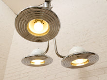 Load image into Gallery viewer, A three-light ceiling fixture with a polished chrome finish and white dome shades, shown illuminated against a white brick wall.
