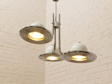Load image into Gallery viewer, A three-light pendant chandelier with white dome-shaped glass shades and polished silver metal rings, shown hanging against a white brick wall.
