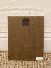 Load image into Gallery viewer, The back of a square brown textured art print or canvas, featuring a metal hanging loop at the top and a small white label in the bottom right corner, leaning against a white brick wall on a woven rug.
